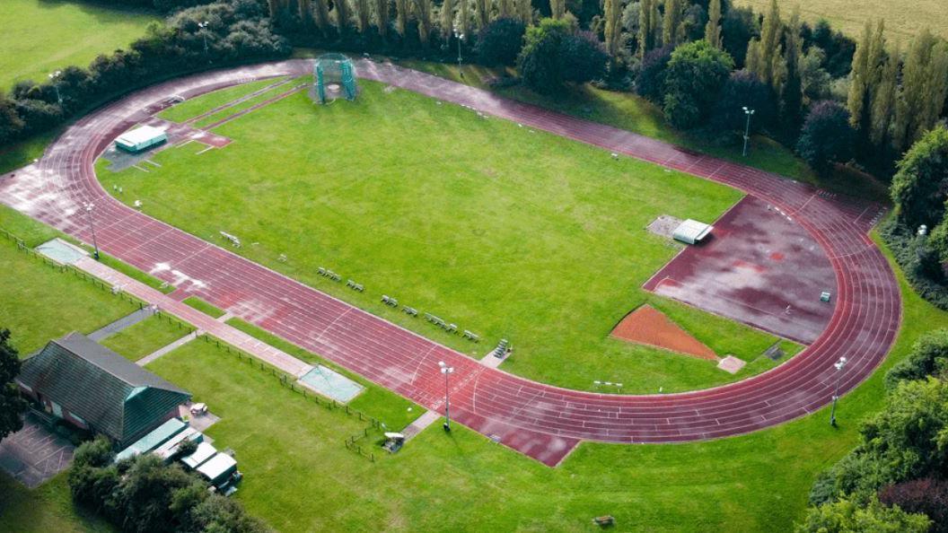 An aerial view of the athletics track shows a green space with a red running track, and trees lining the site. The roof of a green pavilion can be seen on one side. Beyond the track, there are fields and further green spaces.