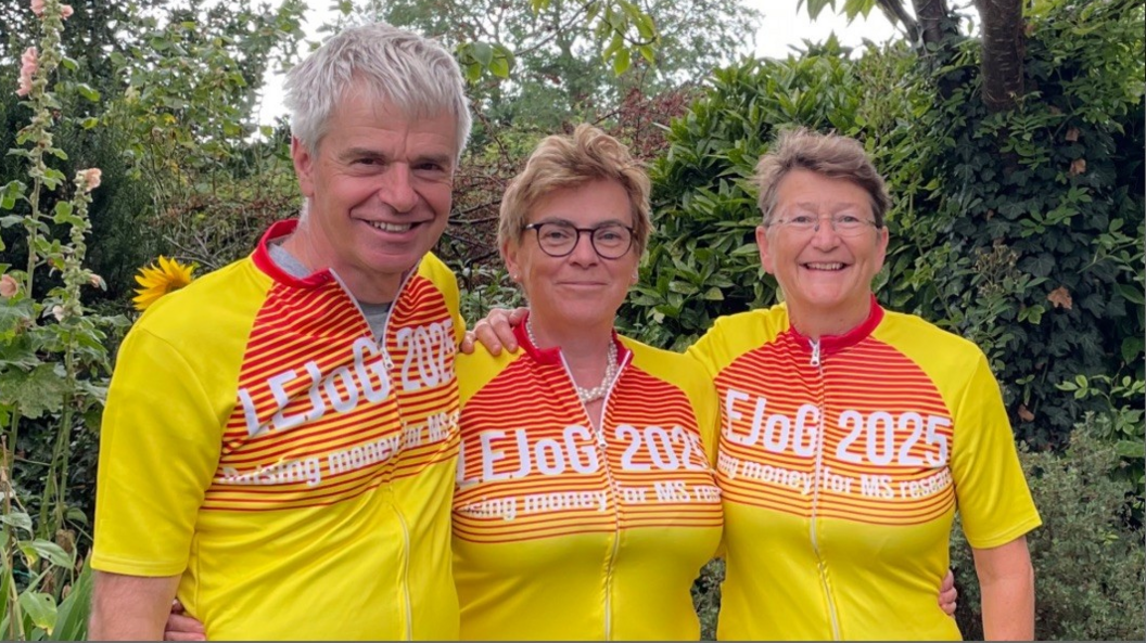 Three people standing arm-in-arm in front of green bushes wearing yellow and orange cycling kit with white writing on it. The man on the left has short grey hair. The woman in the middle has short ginger hair and is wearing glasses. The woman on the right has short light brown hair and is wearing glasses. They are all smiling at the camera.