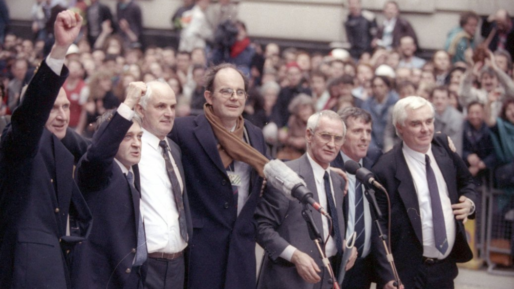 A photograph of seven men in suits standing in front of microphones, with a large crowd in the background. The two men on the right have raised their fists in the air in victory.