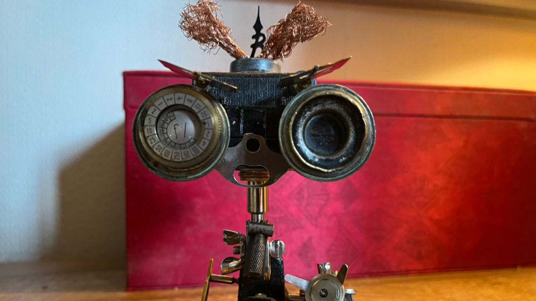 A small robot sculpture, made of metal parts, and one of the two eyes is an old fashioned dial