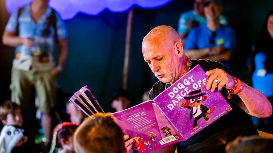 A shaven-headed man with a white goatee beard reads a book titled "Doggy Dance Off" to children in a tent