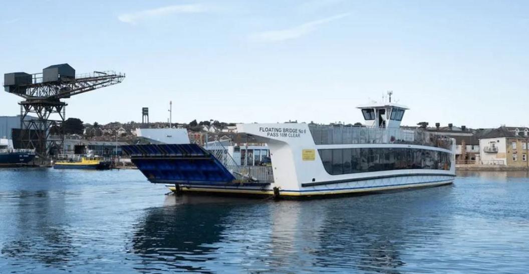White chain ferry mid crossing on a stretch of river. To the right on the shore are rows of buildings to the left of the ferry is a large crane.