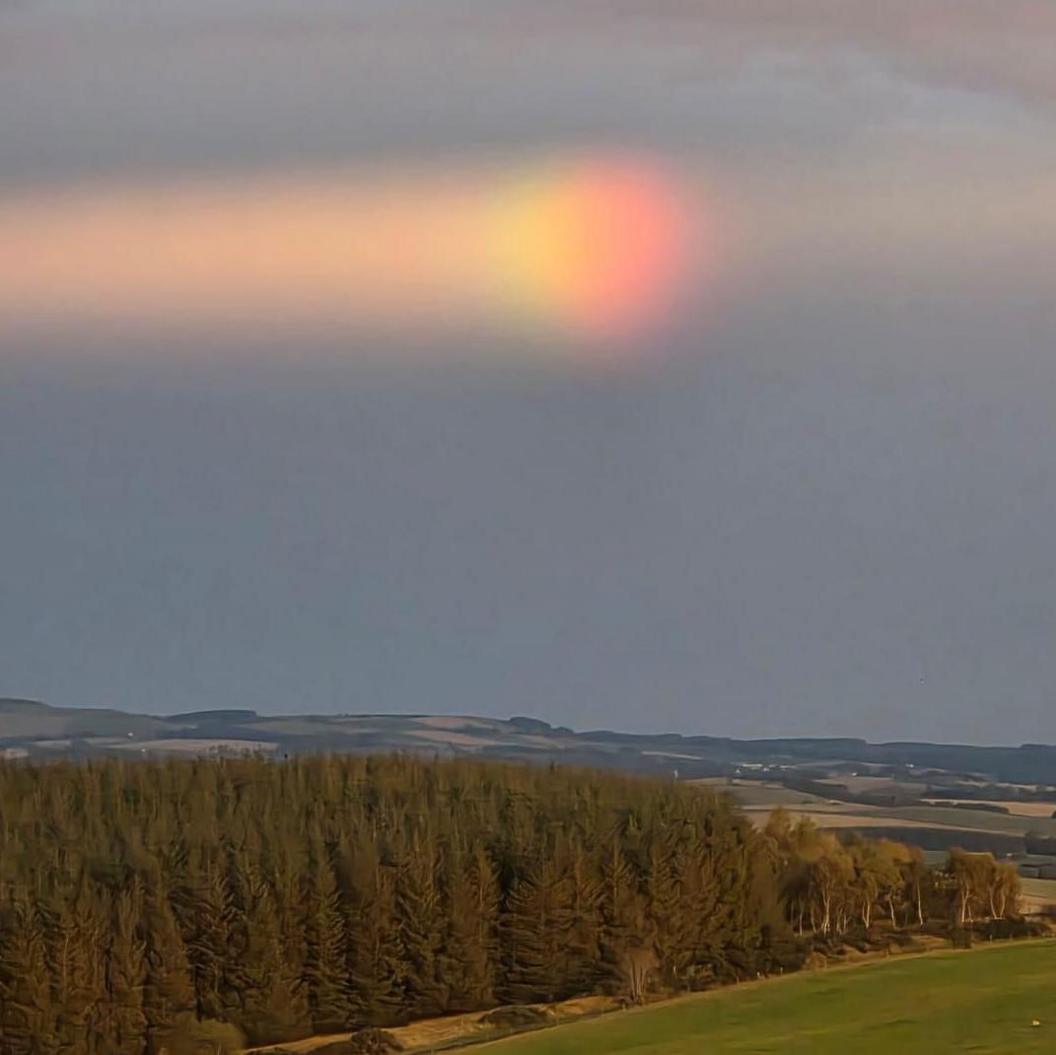 A spectrum of colours appears like a ball leaving a rainbow trail behind it. The spectacle appears over a rural landscape of fields and commercial forestry.