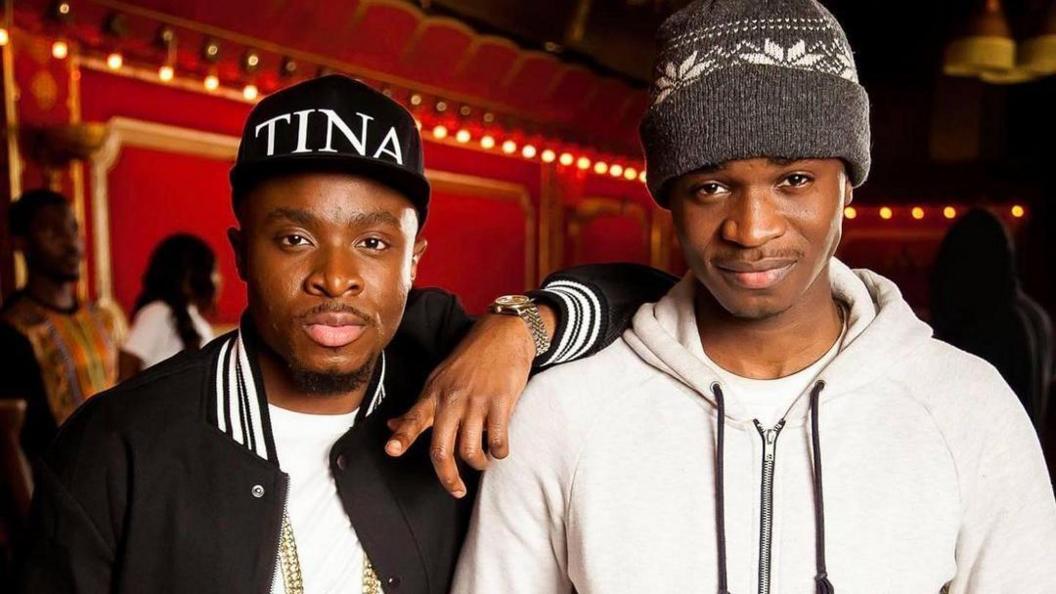 Two young men stand inside a grand red room. The man on the left wears a black baseball cap with the word ‘TINA’ written across it. The one on the right wears a woolly hat and a white hoodie.