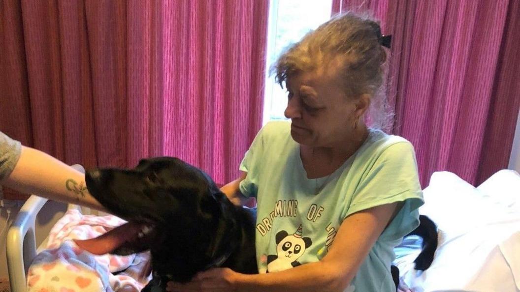 Carol sitting on her bed at the hospice cuddling a black dog, probably a Labrador. She is wearing a green pyjama T-shirt with a cartoon panda on. There are purple/pink curtains behind her and someone's arm is reaching across to stroke the dog, which has its mouth open and tongue out.