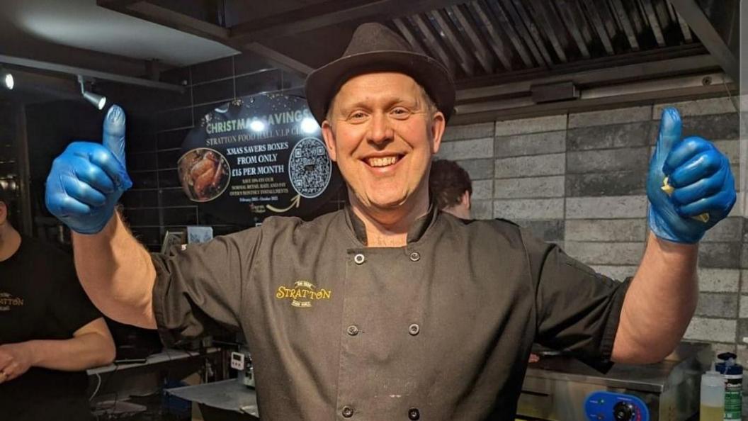 A deli worker with blue gloves and a black trilby hat smiles at the camera