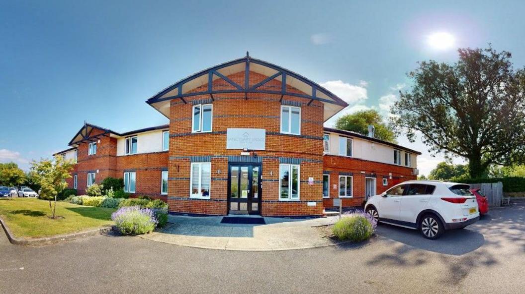 The rehab centre has a new build look, made of new brickwork. It has two levels. The sky is mainly clear and blue. There are cars parked outside and also a small grass area.