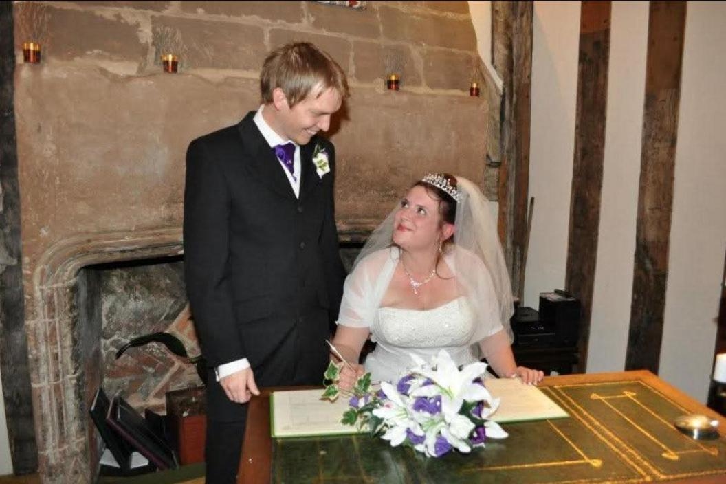 Gavin Hobbins, dressed in a black suit with a purple tie, standing next to his wife, who is sitting by a registry desk in a white wedding dress