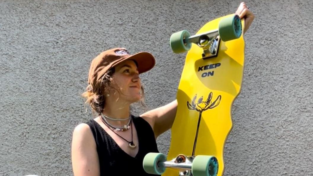 A woman wearing a black, sleeveless top and brown cap is standing in front of a pebble-dash wall. She is holding a bright yellow skateboard that has the message saying Keep On painted on it above a drawing of a flower. The skateboard has kiwi-green wheels.