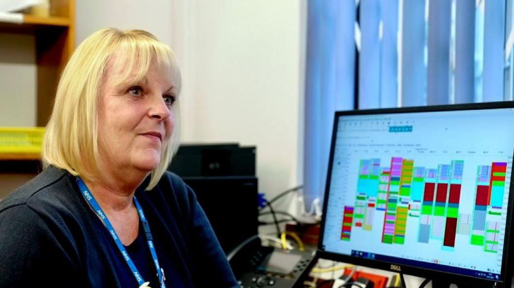 Julie Page sits in front of her computer screen. She has blonde hair in a bob and is wearing a blue top. A blue NHS lanyard is round her neck.