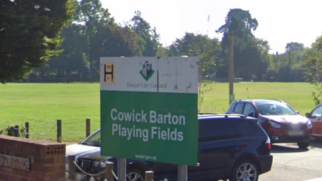 A green and white sign post with the words 'Cowick Barton Playing Fields' at the bottom. At the top, it has the words 'Exeter City Council' with its logo above. Behind the sign are parked cars and a green field. To the left of the sign is a red brick wall.