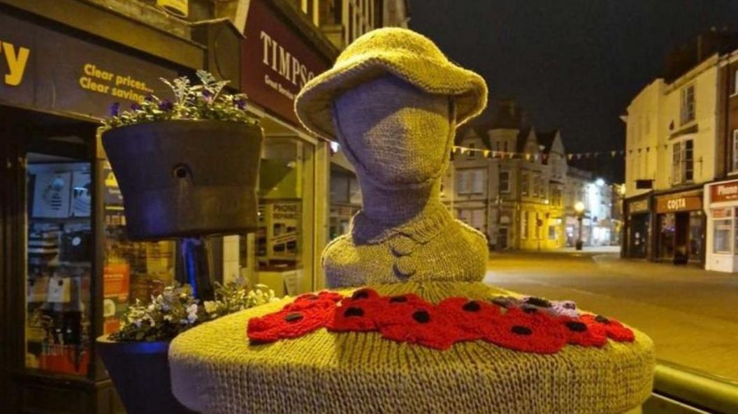 A postbox topper in a town centre with a soldier's helmeted head in grey and red and black poppies underneath it