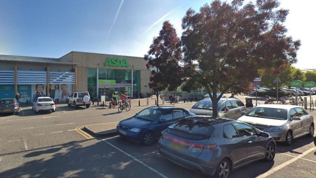 The outside of Asda, showing a large brick building with the Asda logo above the entranceway. In the foreground are parked cars and a tree.