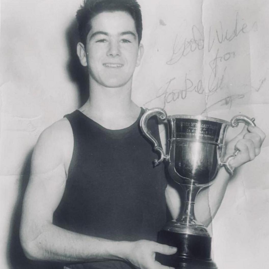A vintage black and white photo of a young Frank Gilfeather wearing a dark tank top holding a trophy