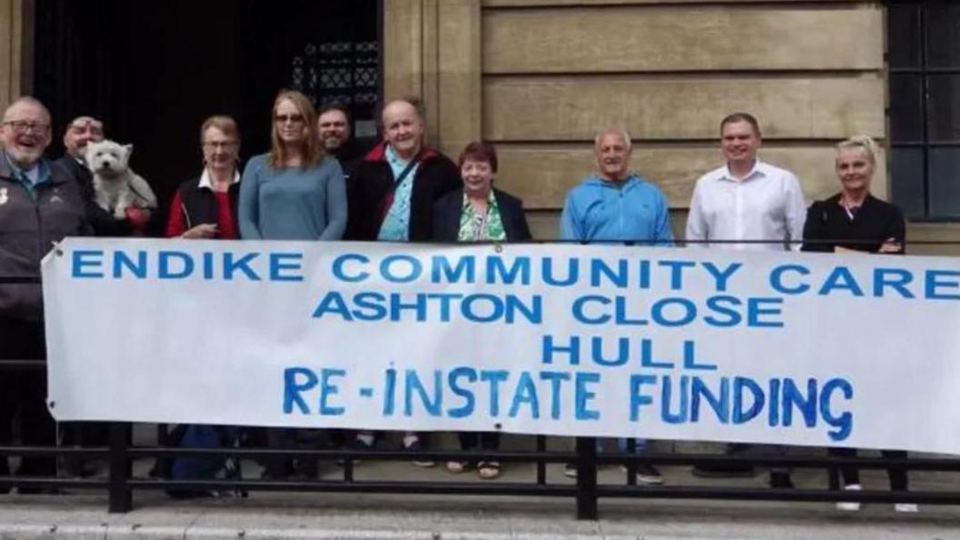 Staff and families in front of Hull's Guidhall behind a large banner stating, 'Endike  Community Care, Ashton Close, Hull, reinstate funding'