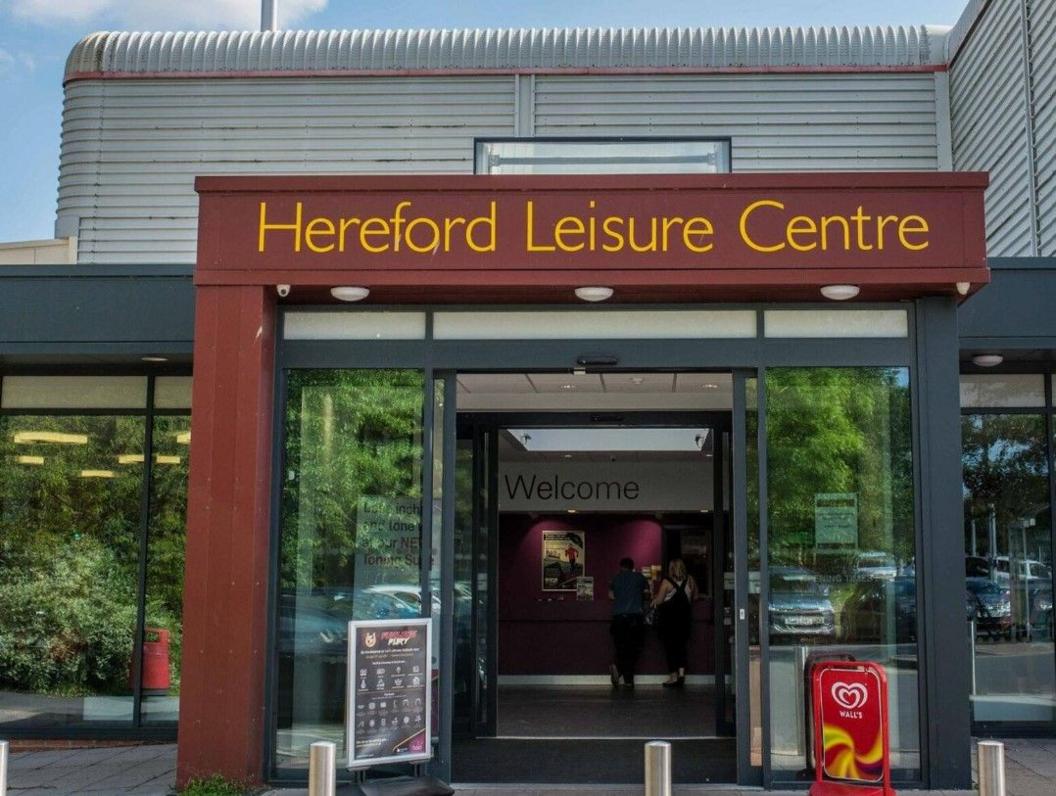 Entrance to Hereford Leisure Centre, with that name being in yellow letters above the door. Blue sky can be seen above the silver building.