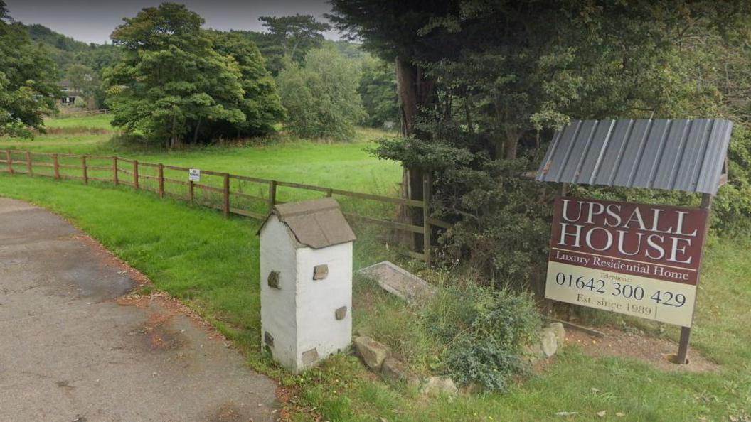 A brown sign that reads 'Upsall House, luxury residential home, Est. since 1989'. A brown wooden fence runs alongside it with a green field behind the fence. A road is to the left of the photo.