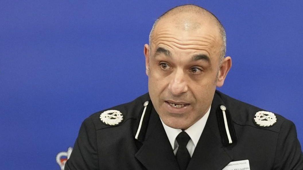 Thom has short cropped grey and black receding hair and brown eyes. He is wearing a black police uniform blazer with crest logo on both shoulders over a white shirt and black tie. He is talking while looking to the left.