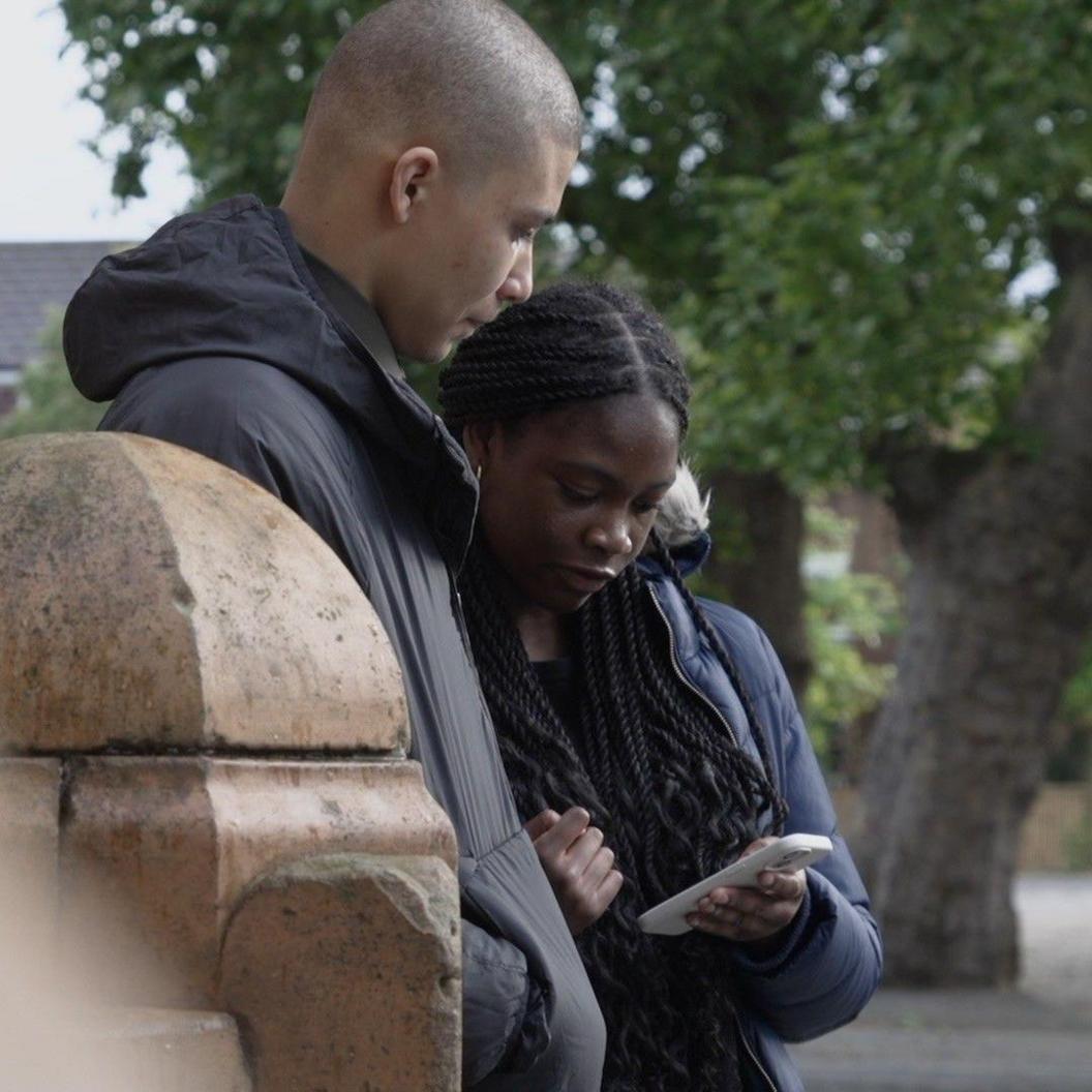 Jason and Tyra, two teenagers, are standing beside a wall on a street, in front of two trees. She is holding a phone in her hand and appears to be showing him it. They are both wearing coats. 