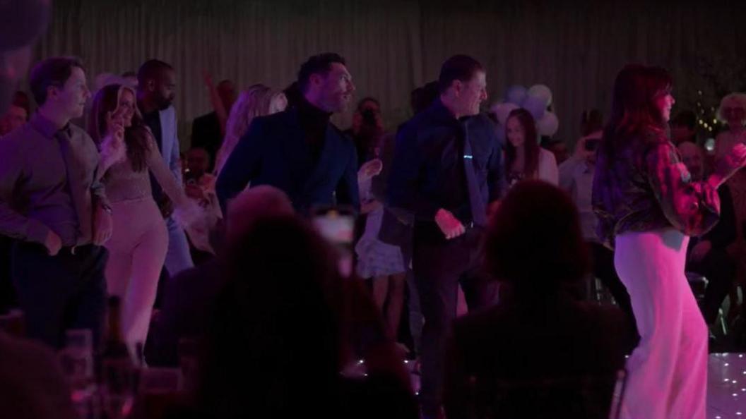 The cast perform a line dance on a dimly-lit dancefloor at a party. They are surrounded by people sitting and standing who are watching them.
