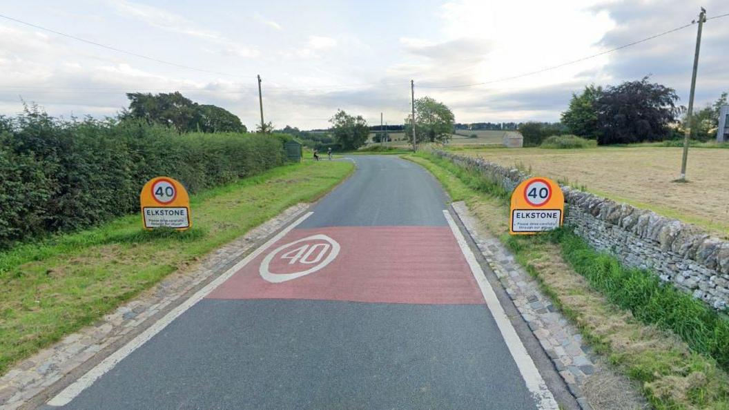 A screenshot from Google street view showing the 40mph signposts when entering Elkstone village. There is a tall hedge on the left of the road, and a low brick wall bordering a field on the right. 