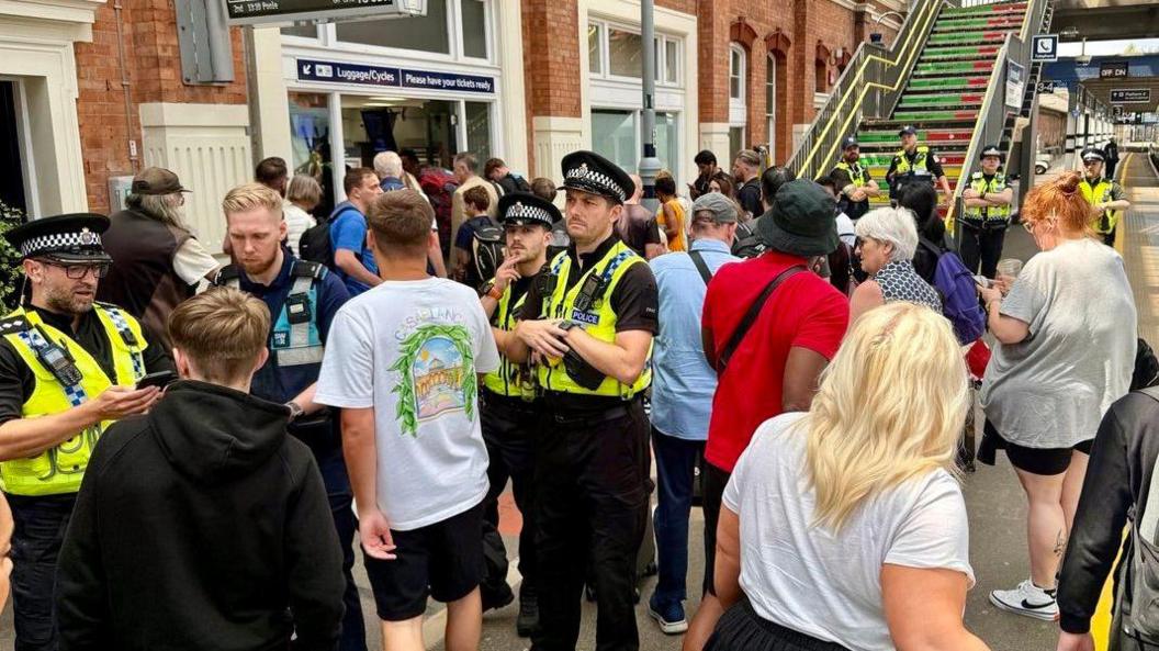 A crowded Bournemouth station platform shows police officers standing and talking to visitors, with many people walking towards the exit.