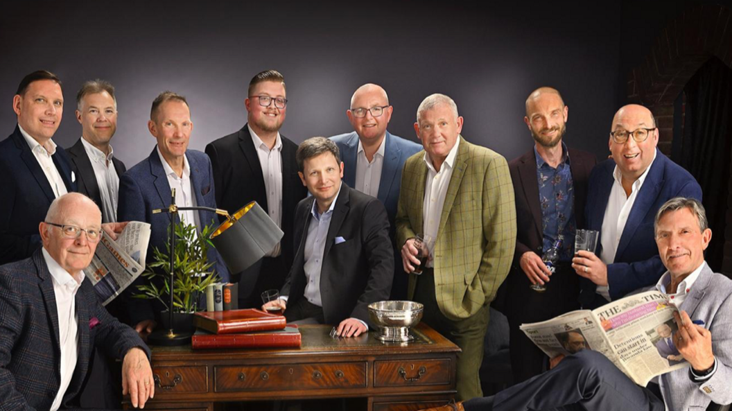 Eleven men, all wearing suits, are ranged round a desk with a lamp and plant on it as well as books. Two of the men are holding newspapers, while three are holding glasses and one holds a bottle.