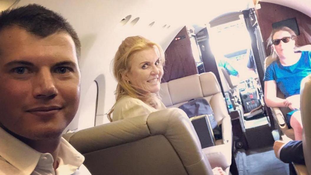 A social media photo showing Sarah Ferguson smiling at the camera in the centre of the image as she sits in what appears to be a small private jet. In foreground is Michael Evers, a young man with brown hair cropped short and wearing a white shirt, open at the neck.