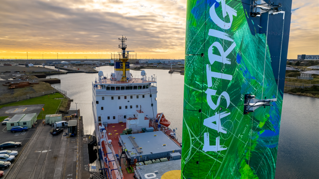 A close-up drone view of the tall rectangular green and blue sail fitted to the deck of the cargo ship. It has the word 'FASTRIG' written vertically on it The ship's funnel and bridge can be seen in the background.