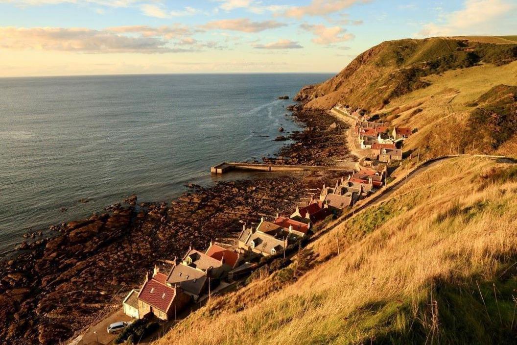 The village's row of traditional houses are below a hillside and above a rocky foreshore at the sea.