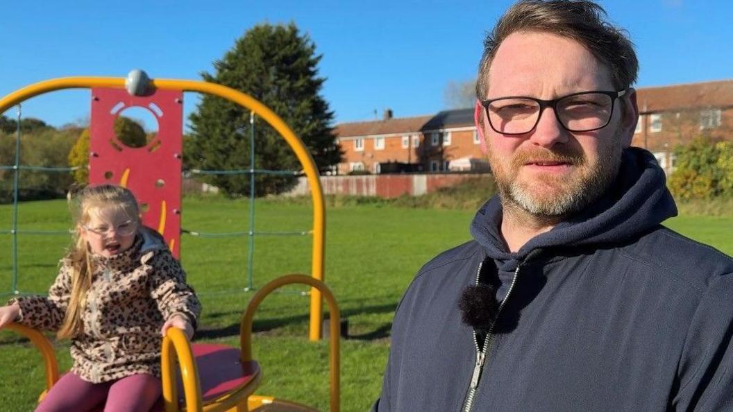A very young girl wearing a floral coat and pink leggings is playing on a metal roundabout in a park while her Dave Armstrong, wearing a blue hooded jumper and spectacles, is looking at the camera. He has short, reddish blond hair and a short beard.