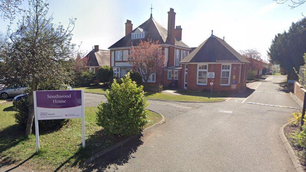 A red brick care home in the background. A grey driveway leads up to it and and there is a white sign in the foreground which states Southwood House. 