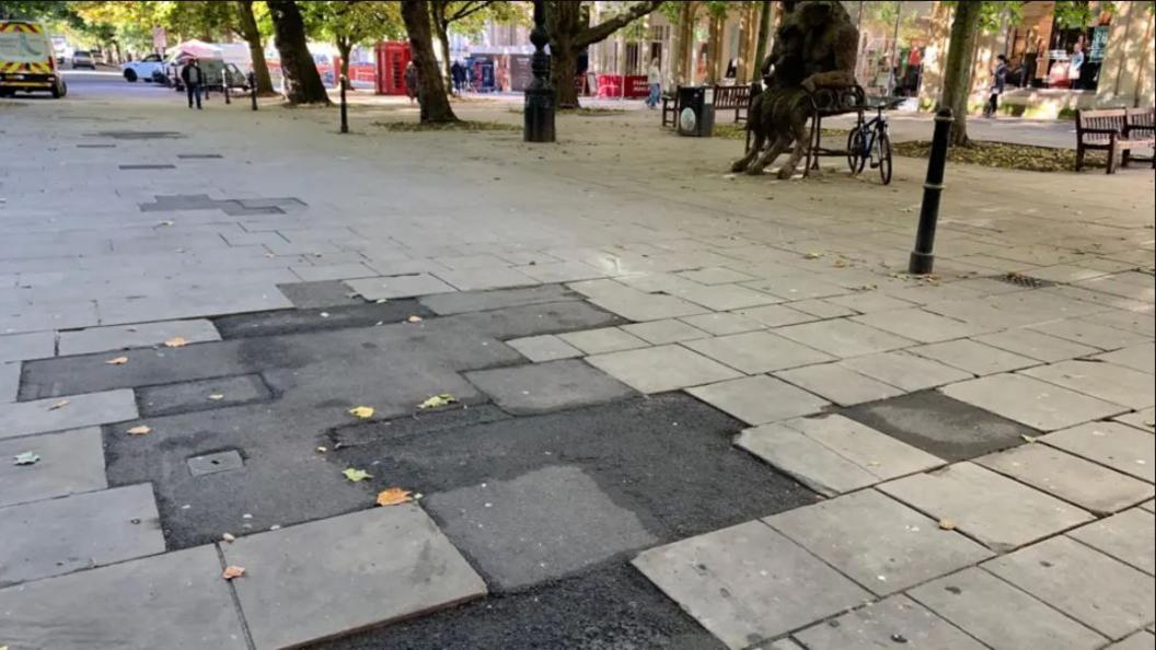 A close-up of the 'tarmac cowpat' on Cheltenham's Promenade. A large patch of asphalt sticks out, surrounded by stone paving slabs. Shoppers can be seen walking in the distance. There are trees lining the Promenade, as well as benches and statues.