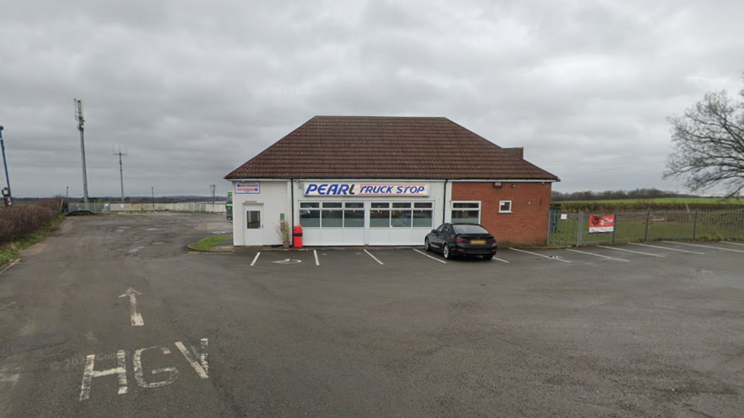 A general view of The Pearl Truck Stop, a small single-storey building with a car parked in front and a HGV sign painted on the road