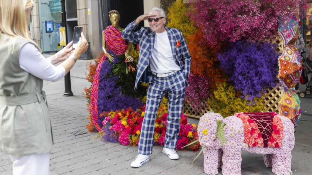 A man in a blue and white suit salutes for his wife alongside an Indian-themed flower installation. 
