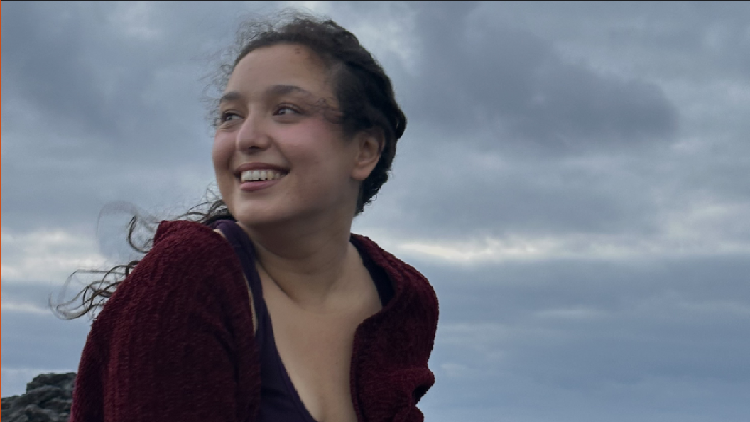 Lara Harris is outside and is looking away from the camera. She is smiling and is wearing a red cardigan. It is cloudy and clearly windy.