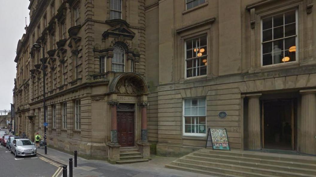 The door to Bolbec Hall, which is has an arched entrance way supported by two pillars. A sign for The Lit and Phil is standing outside the neighbouring building