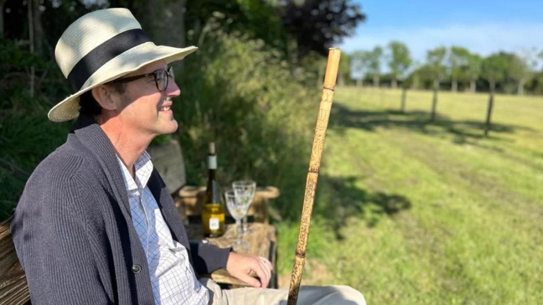 A smiling Simon Boas wearing a panama hat, sitting on a chair beside a green field