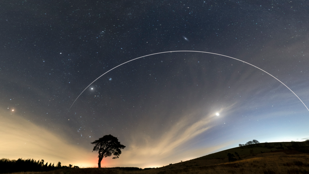 A picture of the night sky. In the foreground are hills and trees.  In the picture you can also see stars and planets.