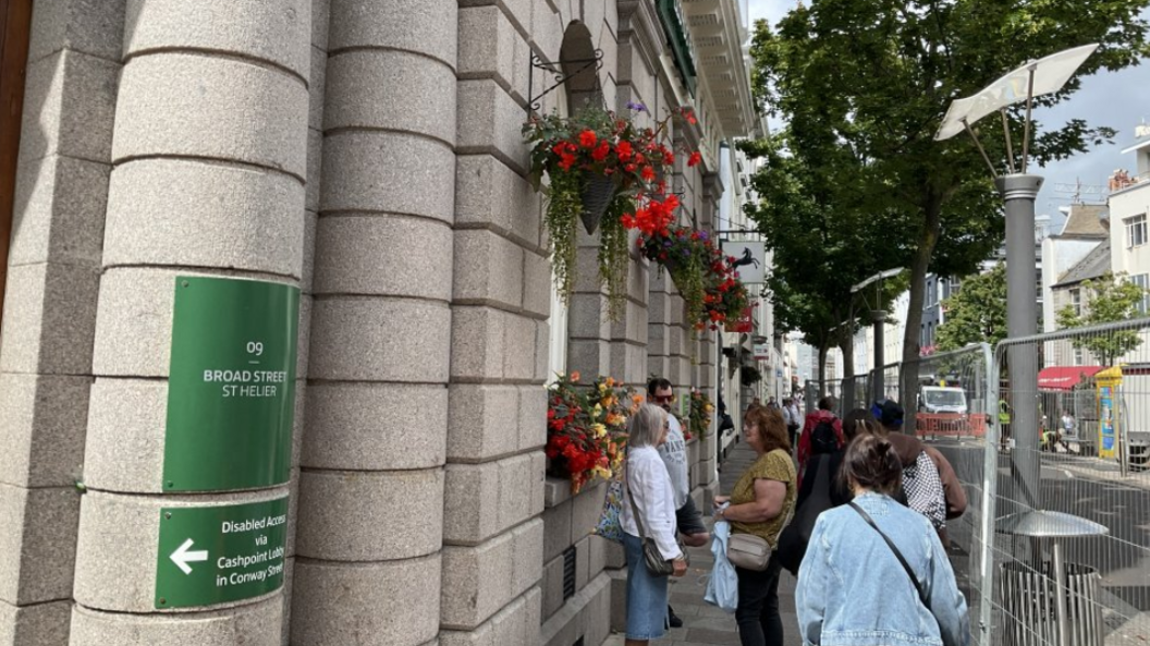 A view of Broad Street which has fencing between the pavement and road. There are people stood on the pavement and there is a building on the left. On the side of a building is a green sign which has the words 'Broad Street St Helier' on it.
On the building there are hanging baskets with red and green flowers, and other plants on the window ledges.