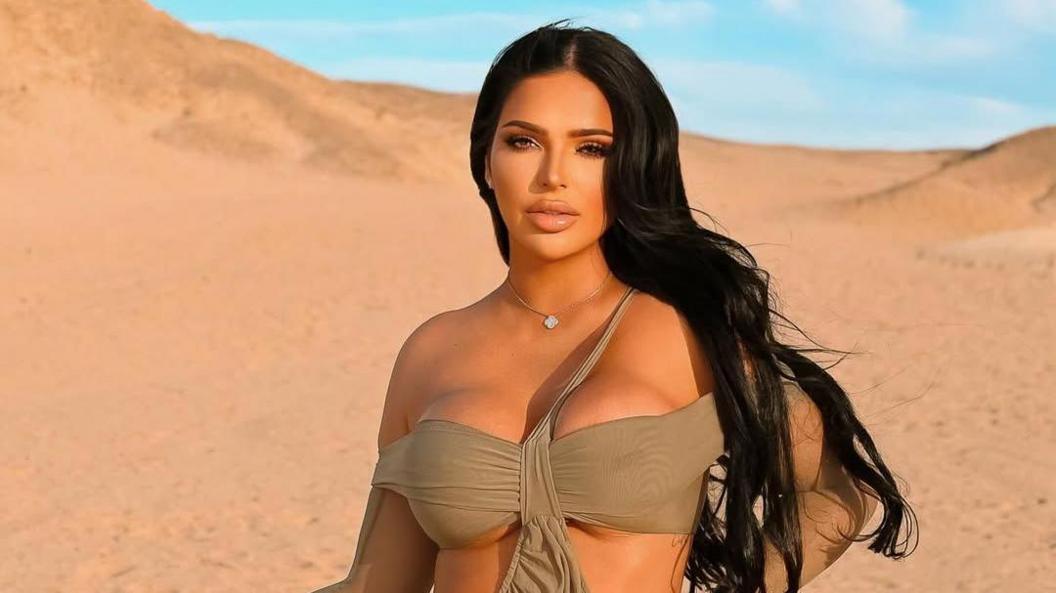 Klaudia Zakrzewska in a desert with blue sky. She wears an off-the-shoulder bikini style top which is taupe. Her hair is long and dark and behind her shoulders. She looks directly at the camera and has a small pendant necklace on. 