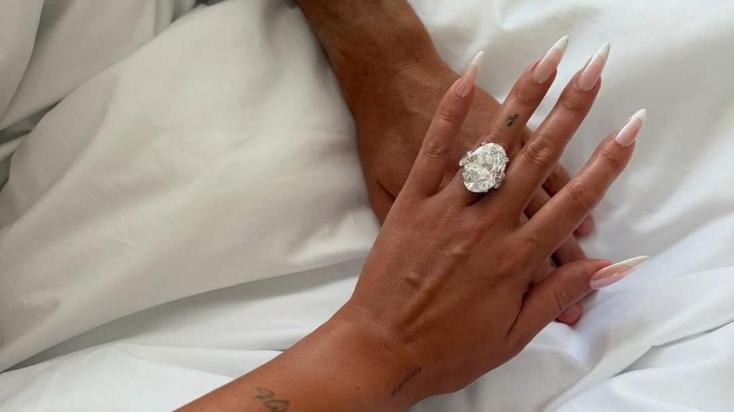Two hands are pictured on top on one another, The top hand has fingers splayed with a long french manicure, and an enormous diamond-style ring on the fourth finger.