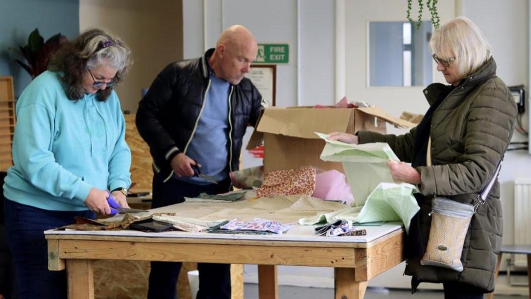 Two women and a man cutting material and looking at designs on a table at a quilt memorial workshop in east Kent in 2025