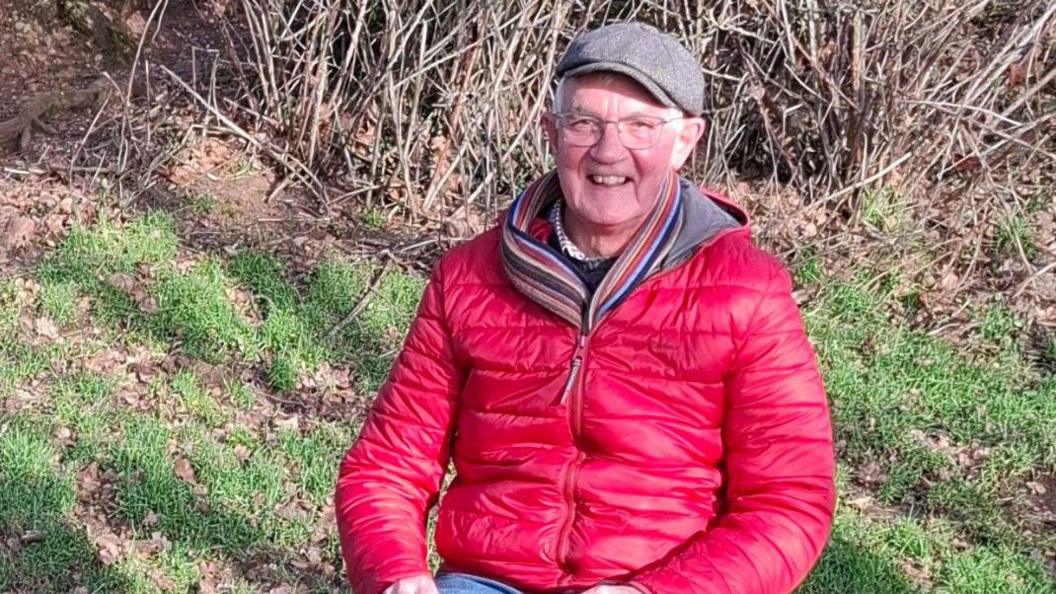 Martin Robinson is wearing a red coat, a scarf of various colours and a cap. He is wearing glasses, smiling and looking at the camera.