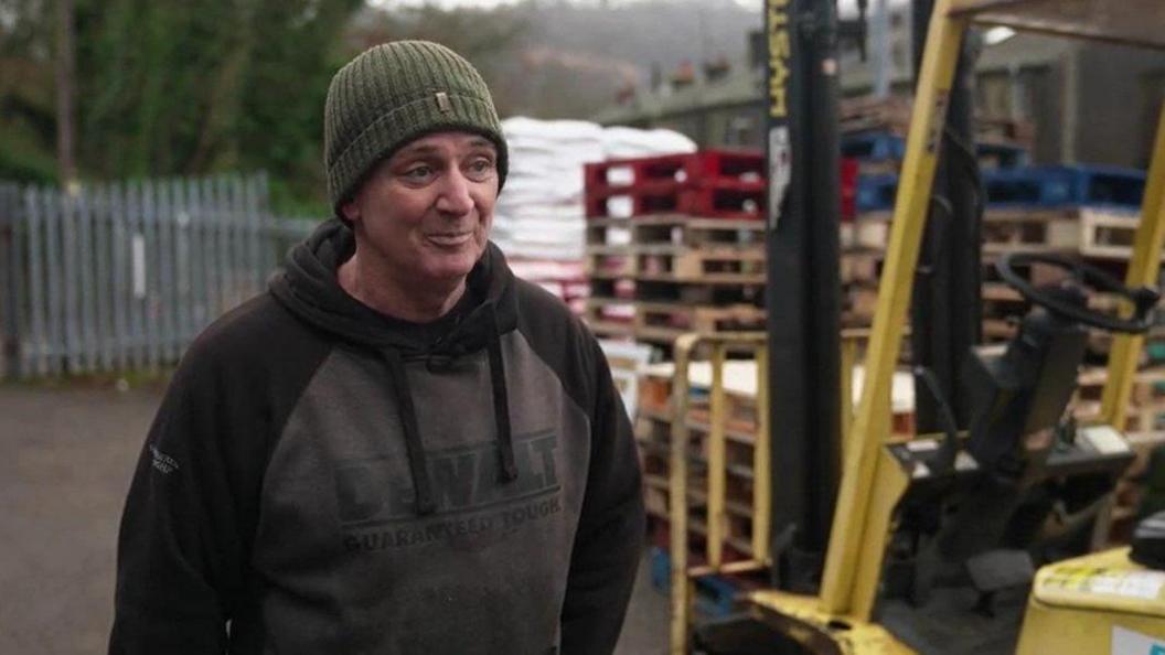 A picture of Paul Jackson who is wearing a green hat and black and grey sweatshirt. He is stood next to a pile of wooden pallets.