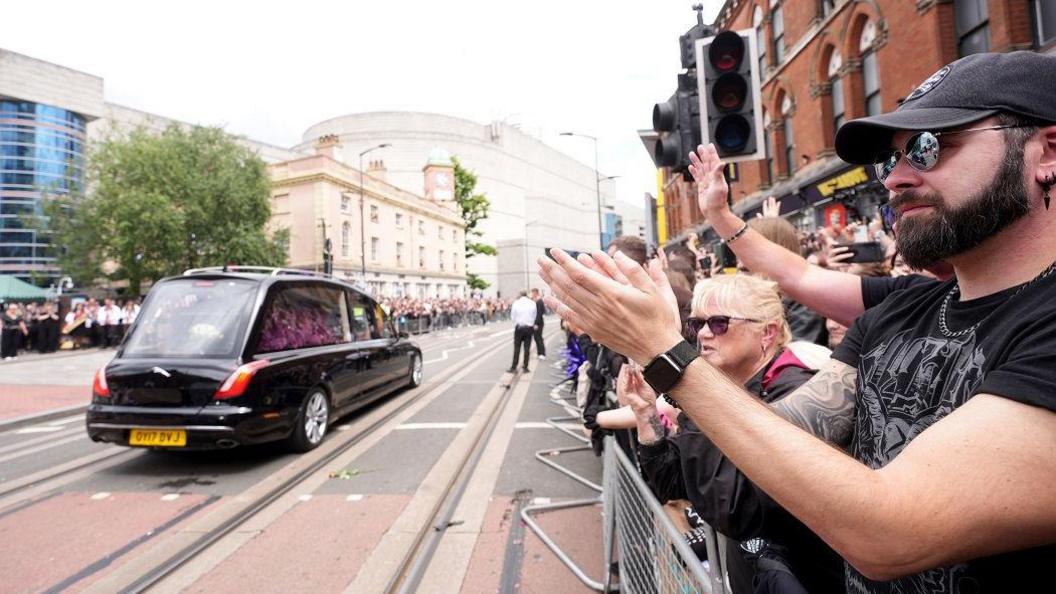 A crowd of people on the right hands side of a street are gathered behind a metal barrier and are clapping. On the road is a black funeral car, inside the window is a display of purple flowers that reads "Ozzy".