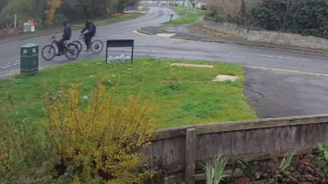 The two teenagers who were later convicted riding bikes past a CCTV camera. They are dressed in all black and are wearing balaclavas to cover their faces. They are driving past a road junction with a grassy patch to their left.