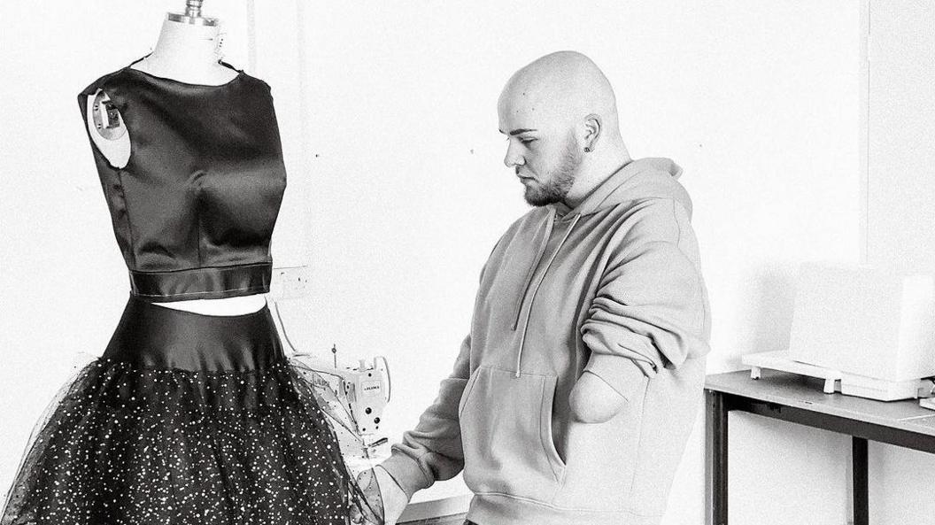 Ryan Rix wearing a hoodie and looking at a clothing stand on which is one of his designs, a black top and skirt