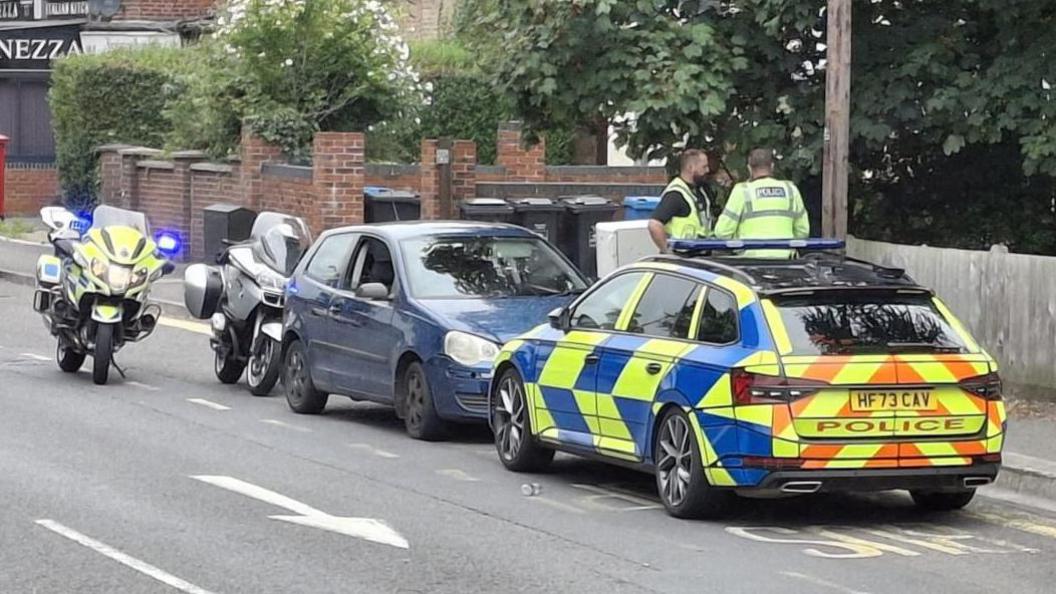 A police car and a police bike parked next to an old blue car and a motorbike at the side of a road. Two police officers are standing next to the vehicles.