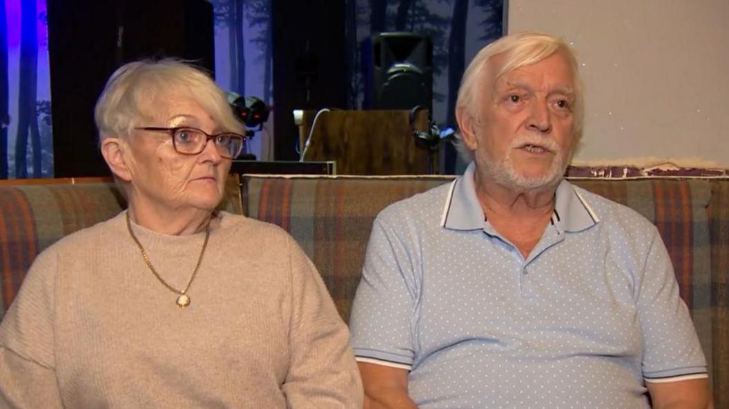 Gill Borg Fenech and Raymond Beeson sit on a sofa in a club. She has short white hair and wears dark-rimmed glasses, a gold pendant necklace and a light-brown jumper. He has white hair and a short white beard and is wearing a light blue shirt with white trimmings. 